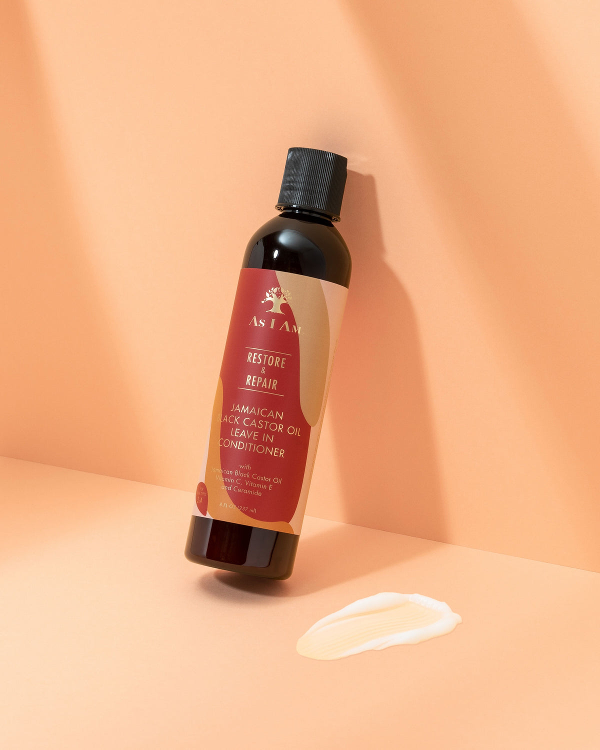 En brun plastikflaske med As I Am Jamaican Black Castor Oil Leave-in Conditioner på en lys overflade. Produktet har en cremet konsistens, som vist i en lille prøve ved siden af flasken.