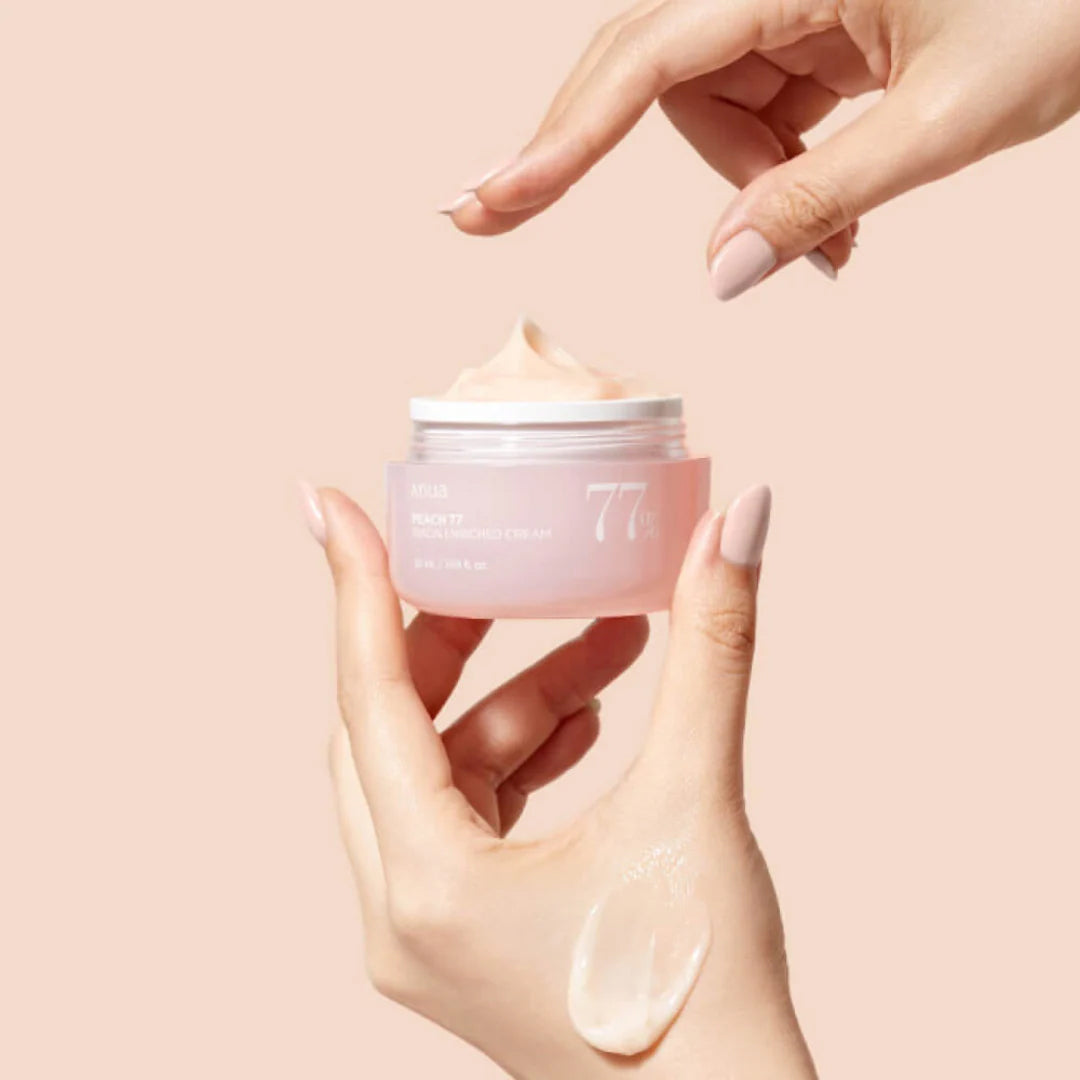 Hånd påfører Anua Peach 77 Niacin Enriched Cream 50ml fra lyserød krukke med cremet ferskenfarvet konsistens.