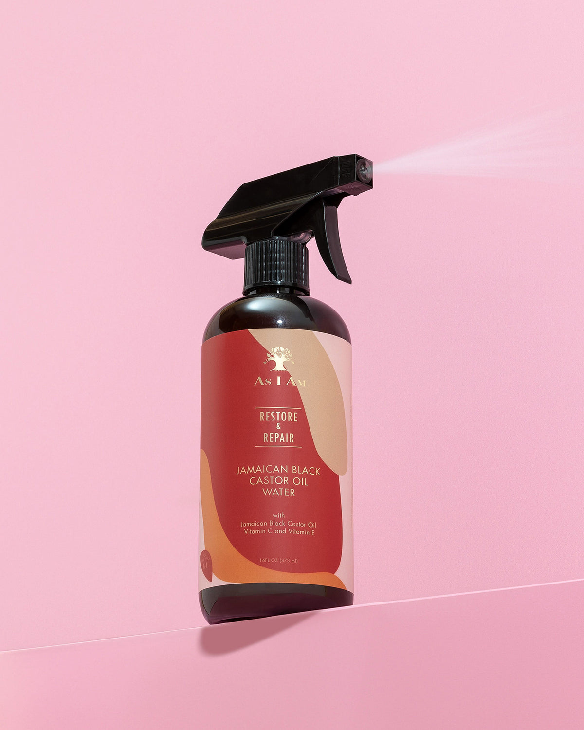 En mørk sprayflaske As I Am Jamaican Black Castor Oil Water (473 ml) sprøjtes mod en lyserød baggrund, der viser en fin tåge af produktet.
