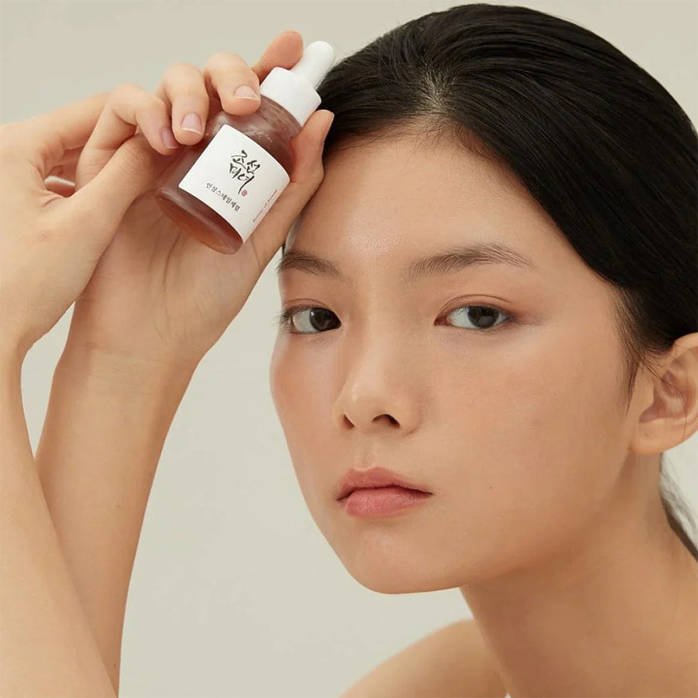 Kvinde med glødende hud holder Beauty of Joseon Revive Serum: Ginseng + Snail Mucin 30ml op mod panden.