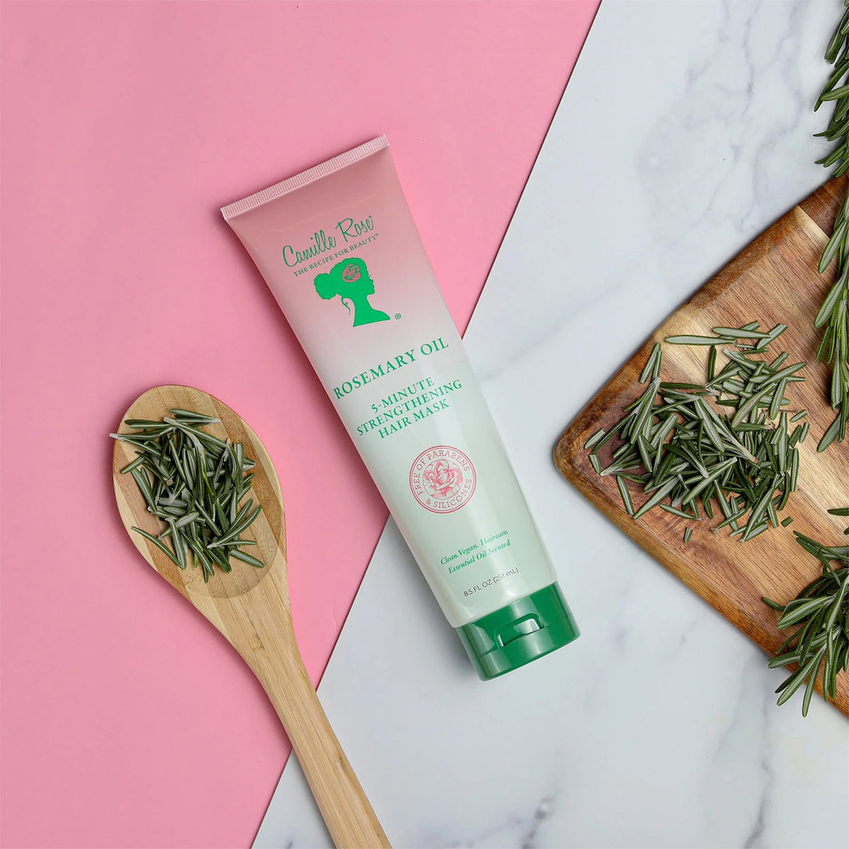 En tube med Camille Rose Rosemary Oil 5-Minute Strengthening Hair Mask ligger på en lyserød baggrund sammen med en træske fyldt med friske rosmarinblade og en skærebræt med yderligere rosmarin.