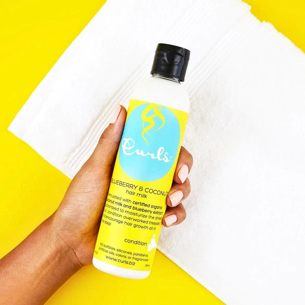 En hånd holder en flaske Curls Blueberry & Coconut Hair Milk mod en hvid baggrund.