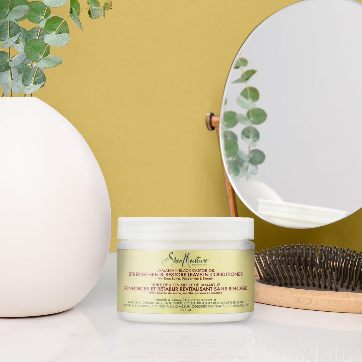 Nærbillede af Shea Moisture leave-in balsam i et lyst badeværelse med et rundt spejl og en træbørste.